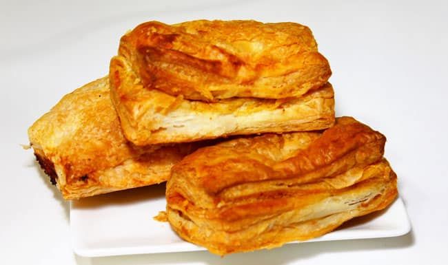 Hand holding a Maruti Live Puff pastry dipped in tomato ketchup with tea service in the background